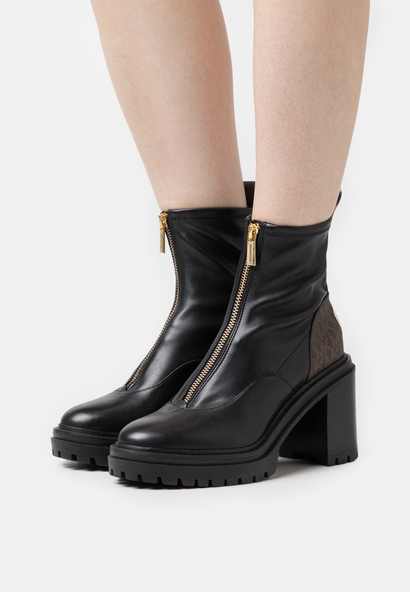 Bottines en cuir noir avec talons épais et fermetures éclair dorées à l'avant, portées sur des jambes nues sur fond blanc.