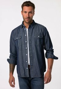 Camisa de mezclilla azul oscuro, de mangas largas, con botones en el frente, dos bolsillos en el pecho con solapas, puños enrollables, llevada sobre una camiseta blanca.