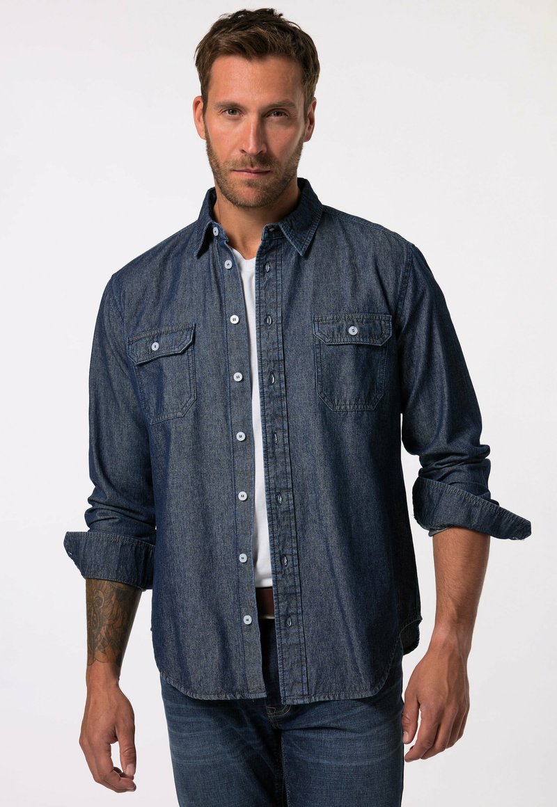 Camisa de mezclilla azul oscuro, de mangas largas, con botones en el frente, dos bolsillos en el pecho con solapas, puños enrollables, llevada sobre una camiseta blanca.