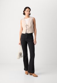Beige mouwloos topje met front-knopen, gecombineerd met zwarte flared jeans. Het model houdt een kleine lichtgekleurde handtas vast en draagt tan kleurige hakken.
