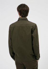 Chaqueta de camisa de algodón verde oliva con collar, cierre de botones y corte recto. La tela tiene un acabado suave y detalles de costura mínimos.