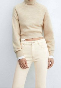 Pull en tricot beige à col roulé avec des poignets et un ourlet côtelés, associé à un jean taille haute jaune clair et une chemise blanche en dessous.