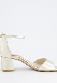 Metalické zlaté sandály s otevřenou špi�čkou, širokým páskem a úzkým páskem kolem kotníku. Blockový podpatek a hladký povrch, navržené pro stabilitu.