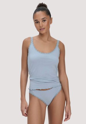 Frau trägt hellblaues, mit Spitze besetztes Camisole und passende Unterwäsche, steht mit neutralem Gesichtsausdruck vor schlichtem Hintergrund.