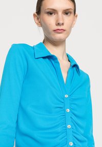 Camisa botonada de riscas em azul claro, com gola, textura franzida na parte da frente e pequenos botões iguais.