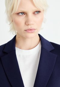 Blazer blu navy con rever strutturati, abbinato a una maglietta bianca a girocollo. Tessuto morbido con un design minimalista e senza motivi visibili.
