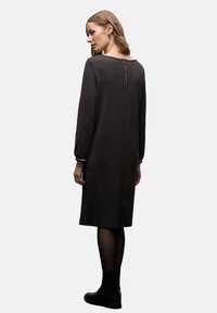 Tummanharmaa pitkähihaisen mekko suoralla siluetilla, pyöreällä kauluksella ja takana vetoketjulla. Siinä on kerätyt hihat ja sileä rakenne.