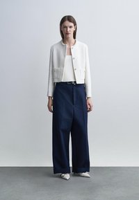 Woman wearing a white cropped textured jacket, white top, wide navy trousers, and white pointed shoes standing on a gray floor.