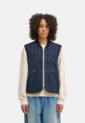 Jeune personne aux cheveux bouclés portant un gilet zippé rayé bleu sur une chemise beige à manches longues et un jean bleu clair, debout face à l'avant.