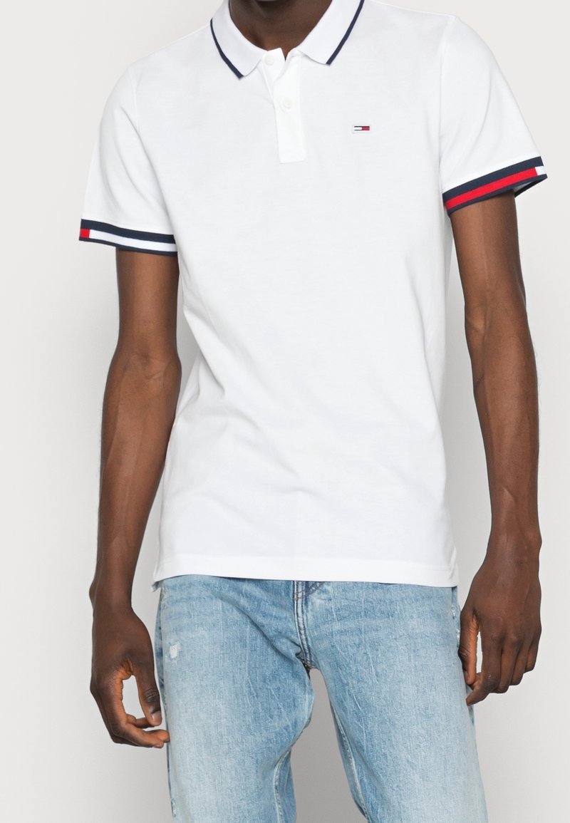 Homme portant un polo blanc à manches courtes avec des bordures de manches rayées bleu marine et rouge, et un jean denim bleu clair.