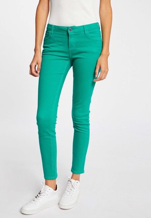 Schmal geschnittene grüne Hose, getragen von einer stehenden Person, kombiniert mit weißen Sneakers und einem in die Hose gesteckten weißen ärmellosen Top.