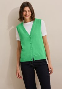 Grön stickad väst, V-ringad design, fem knappar, ribbad nederkant, bärs över en vit t-shirt, ihop med mörka jeans, slät textur.