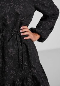 Svart klänning med blommig prägel som har långa ärmar, ett draperat midjeband med snörning och en flerlagerskjol.