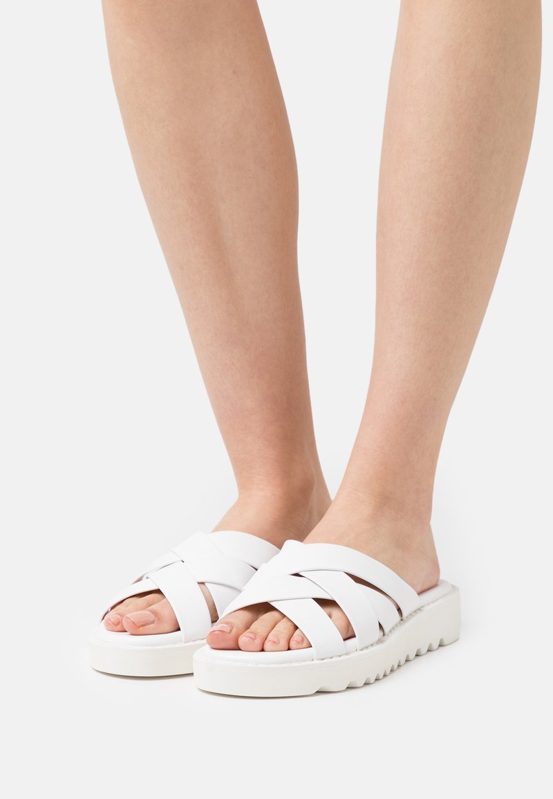 Pieds nus portant des sandales blanches à enfiler avec des brides épaisses croisées et des semelles rainurées sur un fond uni.