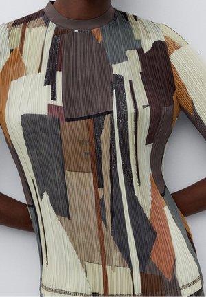 Gros plan d'une personne portant un haut plissé à manches longues avec un motif géométrique abstrait dans des tons marron, beige, gris et orange.