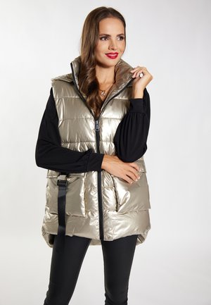 Gilet puffy in oro metallico con colletto alto, dotato di zip frontale, tasche laterali e dettagli con cinturino nero regolabile, indossato sopra un top nero.