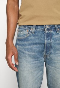 Man i ljusblå slitna denimjeans och en beige skjorta, med en hand avslappnat vid sin sida mot en vit bakgrund.