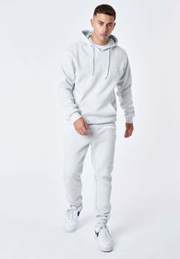 Felpa con cappuccio grigio chiaro e pantaloni jogger abbinati, entrambi in tessuto morbido con una vestibilità rilassata. Presentano una tasca frontale e cappuccio con coulisse. Indossati con scarpe da ginnastica bianche.