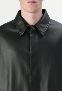 Schwarze Lederjacke mit spitzem Kragen, glatter Textur und einem Reißverschluss auf der Vorderseite. Minimalistisches Design ohne sichtbare Muster oder Beschläge.