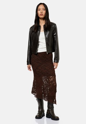 Femme aux longs cheveux noirs, portant une veste en cuir noire, un haut blanc, une jupe en dentelle marron et des bottes noires, debout avec une expression neutre.