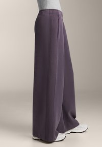 Fialové kalhoty s širokými nohavicemi, texturovaným pasem a bočními kapsami, vyrobené z měkké látky. V kombinaci s bílými sportovními botami.