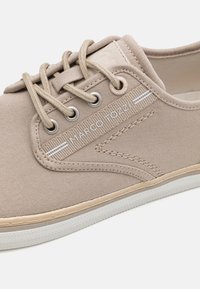 Sneaker in tela beige con occhielli bianchi, lacci beige e suola in gomma testurizzata. Include il logo "Marco Tozzi" e dettagli di cuciture sottili.