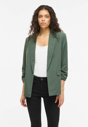 Göndör hajú nő zöld blézerben, fehér felsővel és fekete farmerral, egyszínű fehér háttér előtt állva.