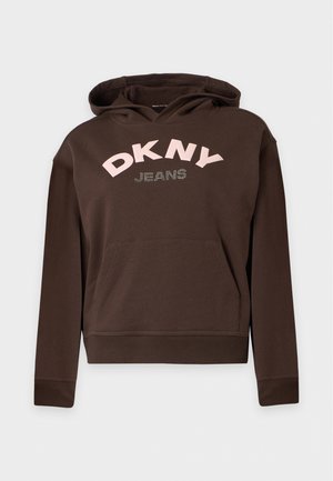 Жена с черна коса, носеща кафяв суичър DKNY, светлосини дънки с широки крачоли и черни обувки на равно ходило, стояща на едноцветен фон.