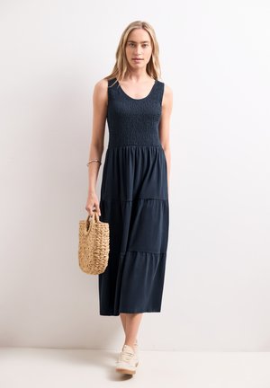 Frau trägt ein ärmelloses, marineblaues Midikleid, hält eine geflochtene Strohtasche und trägt weiße Sneakers, steht vor einem schlichten Hintergrund.