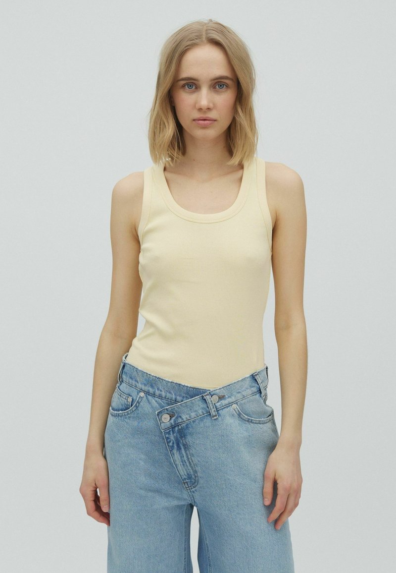 Jeune femme aux cheveux blonds portant un top sans manches jaune pâle et un jean bleu clair avec une ceinture asymétrique superposée.