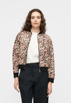 Kvinde med krøllet hår iført en pink bomberjakke med blomsterprint, hvid skjorte og sorte bukser, stående mod en ensfarvet lys baggrund.