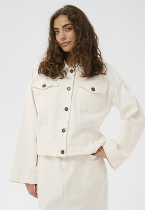 Tânără cu păr lung și creț, purtând o jachetă cu nasturi de culoare alb-închis și pantaloni asortați, stând cu o mână pe șold.