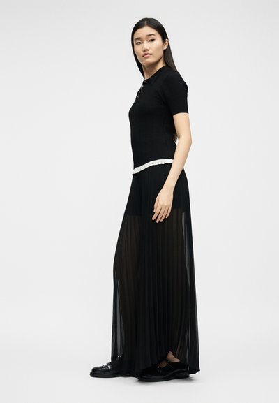 Γυναίκα που φοράει μαύρο πλεκτό τοπ με κοντά μανίκια και μακριά διάφανη πιε ντεσέ μαύρη φούστα με μαύρα παπούτσια, στέκεται μπροστά σε απλό φόντο.