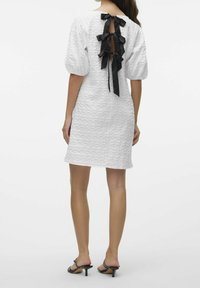 Valkoinen mekko, jossa on teksturoitu kangas, lyhyet puffiset hihat ja avainaukkotakaosa, joka on koristeltu kolmella mustalla satiinikirjalla. Polvenmittainen ja istuva.