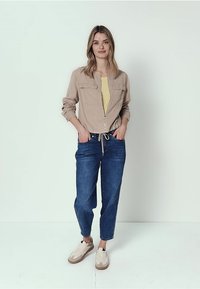 Mujer de pie con las manos en los bolsillos, vistiendo una chaqueta beige con cremallera, camiseta amarilla, vaqueros azules y zapatillas blancas, sobre un fondo claro y sencillo.