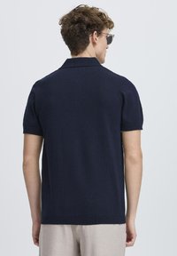 Polo shirt navy cu mânecă scurtă, cu textură țesută, guler și tiv cu nervuri. Modelul poartă pantaloni deschiși la culoare și ochelari de soare, văzut din spate.
