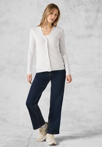 Rippstrick-weißer Cardigan mit V-Ausschnitt und Knöpfen vorne, kombiniert mit dunkelblauen weiten Jeans und beigen Sneakers. Glatte Textur, lässiges Design.