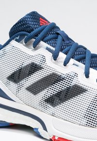 Scarpa sportiva bianca con accenti blu e rossi, dotata di una tomaia in rete, superficie testurizzata e tre strisce nere sul lato.