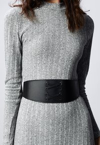 Large ceinture en simili cuir noir avec un détail à lacets à l'avant, portée par-dessus une robe moulante en tricot gris côtelé. Texture lisse, design structuré.