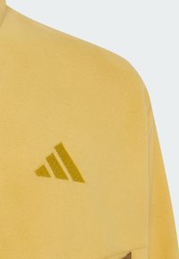 Κίτρινο ύφασμα fleece με μικρό κεντητό λογότυπο τριών κεκλιμένων ραβδώσεων σε πιο σκούρο κίτρινο στην περιοχή του στήθους.