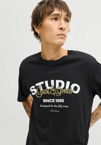 Czarny bawełniany t-shirt z wyraźnym białym i złotym napisem, w tym "STUDIO" oraz "Jack & Jones", o luźnym kroju i okrągłym dekolcie.