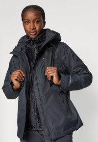 Zwarte geïsoleerde jas met een capuchon en fleece voering, met een ritssluiting aan de voorkant en twee zijzakken. Bevestigde banden op de achtergrond.