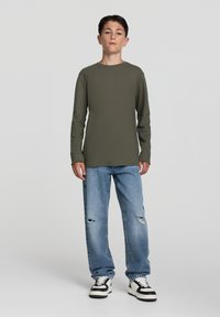 Camicia verde oliva a maniche lunghe abbinata a jeans chiari strappati e sneakers nere e bianche. Outfit semplice e casual con una vestibilità rilassata.