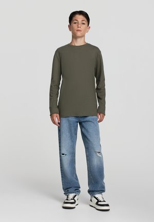 Camicia verde oliva a maniche lunghe abbinata a jeans chiari strappati e sneakers nere e bianche. Outfit semplice e casual con una vestibilità rilassata.