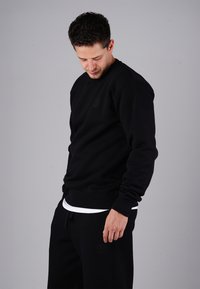 Black sweatshirt with a round neckline, ribbed cuffs, and a small logo. Soft texture, worn over a white shirt, paired with black pants.
