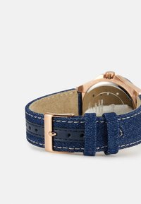 Blå denim klockrem med en ljusbrun innerfoder och en roséguld metallspänne. Remmen har kontrasterande sömmar och hål för justering.
