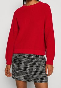 Maglione rosso lavorato a maglia con maniche larghe, scollo rotondo, tessuto testurizzato, abbinato a una gonna a fantasia bianca e nera.