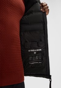 Interno di giacca imbottita nera con una tasca che mostra le etichette di cura, con il logo "STRELLSON". La parte esterna è elegante con chiusura a zip.