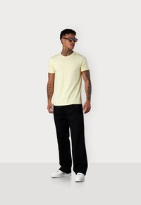 T-shirt en coton jaune avec un col rond, associé à un pantalon large noir et des baskets blanches. Le t-shirt comporte un petit logo.