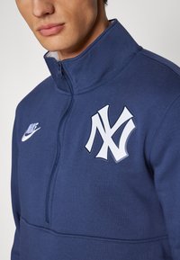 Námornícky modrý mikina s štvrťovým zipsom, s bielym logom Nike a emblémom New York Yankees. Vyrobená z mäkkého materiálu, s hladkou textúrou a prešívanými detailmi.
